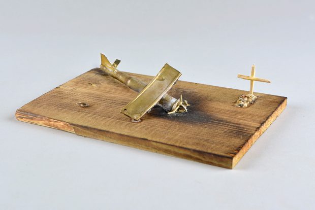 Modèle d’un accident d’avion, sur une planche de bois. L’avion écrasé couvre une bonne partie de la planche, et une tombe, faite d’un amas de pierre avec une croix, se trouve en bordure.