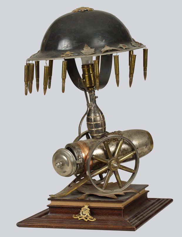 Étrange lampe de table faite de divers objets militaires. L’abat-jour est un casque de soldat autour duquel pendent des cartouches de carabine; la lampe elle-même est faite de douilles de différents calibres.
