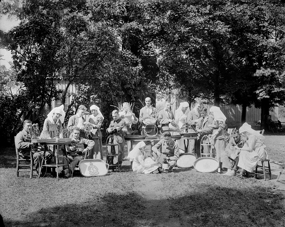 Photo en noir et blanc de soldats de la Première Guerre mondiale en convalescence, accompagnés de leurs infirmières. Ils sont à l’extérieur, par une journée ensoleillée, autour de tables avec des objets d’artisanat.