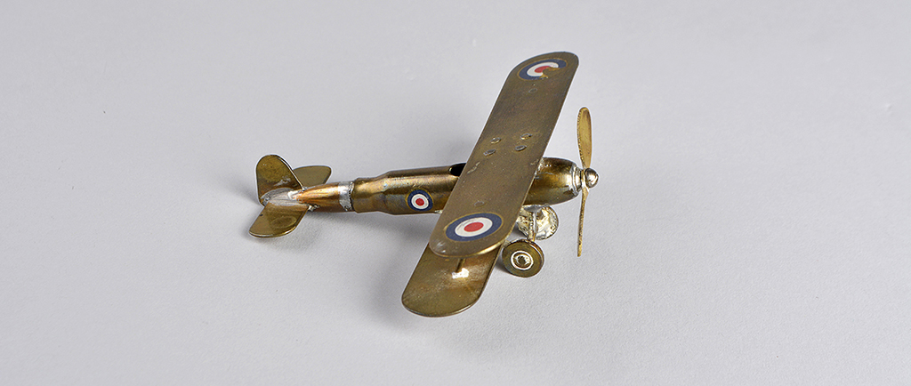 Petit avion en laiton lustré fait avec une cartouche de carabine. L’avion possède une hélice à l’avant et des cocardes sur chaque aile et sur le fuselage.