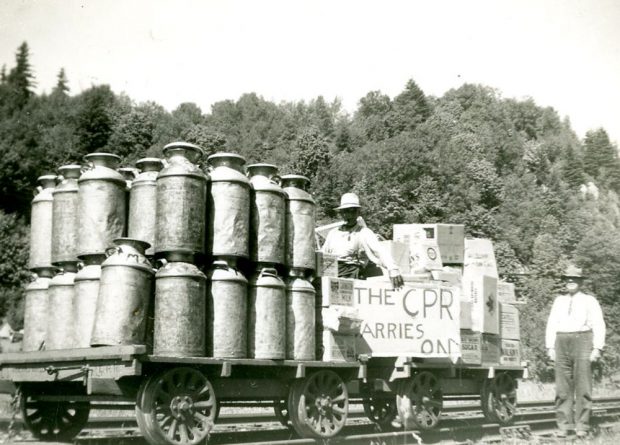 Photo en noir et blanc avec des bidons de lait sur un wagon plat surbaissé. Deux hommes tiennent une pancarte sur laquelle on peut lire « The CPR Carries On » (Le CFCP tient le coup).