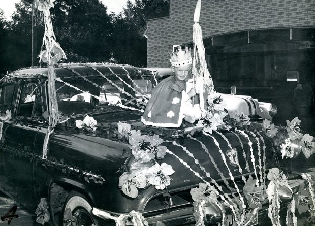 Photo en noir et blanc d'un jeune garçon portant un manteau royal et une couronne, assis sur le capot d'une voiture décorée pour le défilé.