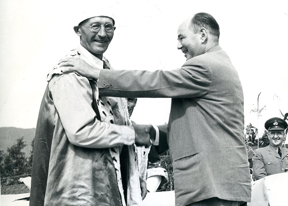 Photo en noir et blanc de deux hommes debout sur un podium qui se serrent la main. Un homme porte un manteau royal et une couronne tandis que l'autre porte un habit, 1954.