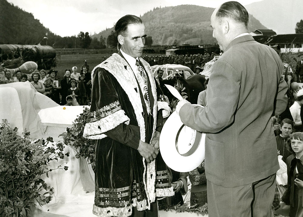 Photo en noir et blanc de deux hommes debout sur un podium. L'un porte un manteau royal alors que l'autre porte un habit et tient un chèque, 1953.