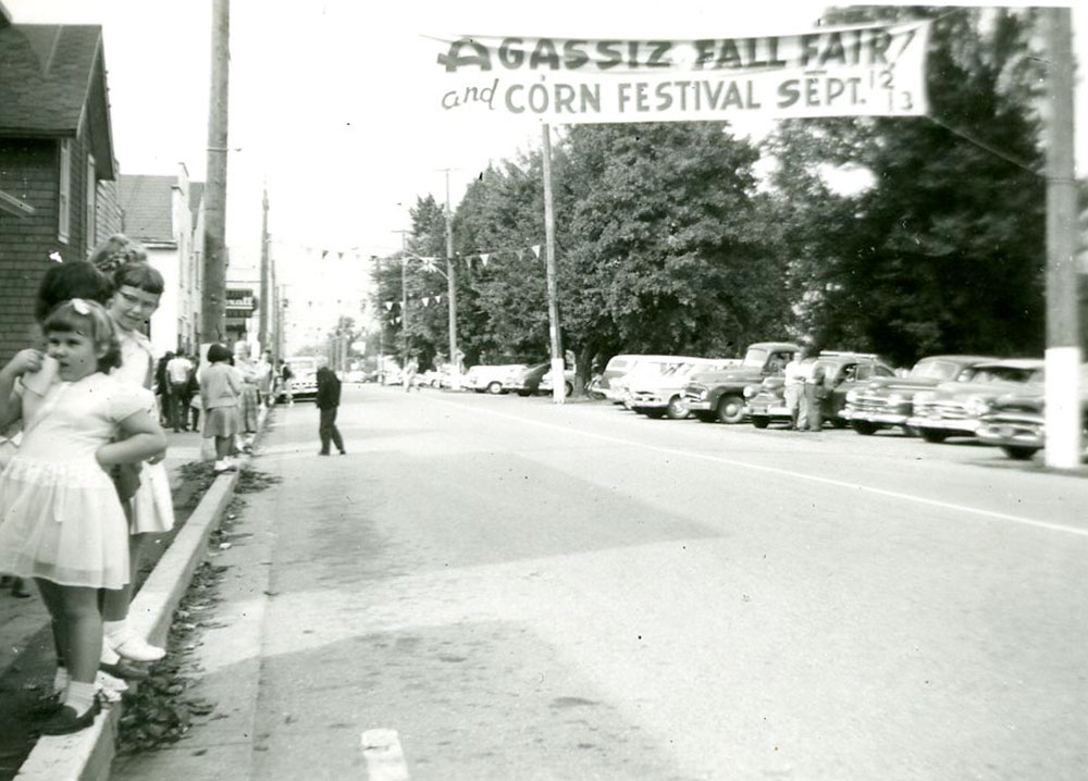 Photo en noir et blanc avec des gens rassemblés au bord d'une rue pour voir un défilé. Sur une bannière qui flotte au-dessus de la rue on peut lire : « Agassiz Fall Fair and Corn Festival », 12 et 13 septembre.