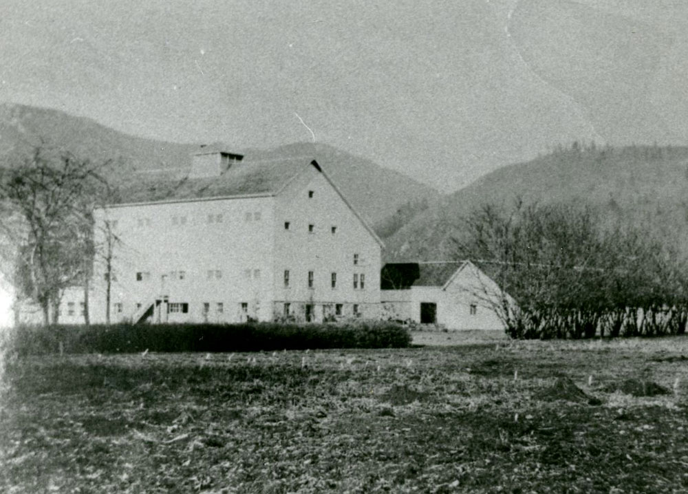 Photo en noir et blanc d'une grange en pierres entourées de champs et de montagnes.