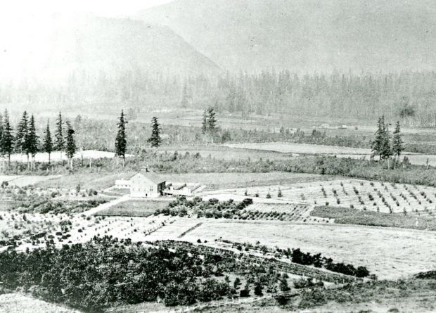 Photo en noir et blanc de la Ferme expérimentale du Dominion d'Agassiz, entourée de champs, d'arbres et de montagnes.