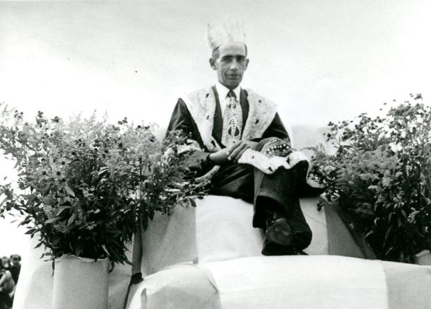 Photo en noir et blanc d'un homme vêtu d'une robe royale et d'une couronne, assis sur un trône et entouré de fleurs. C'est le roi du maïs de 1951.