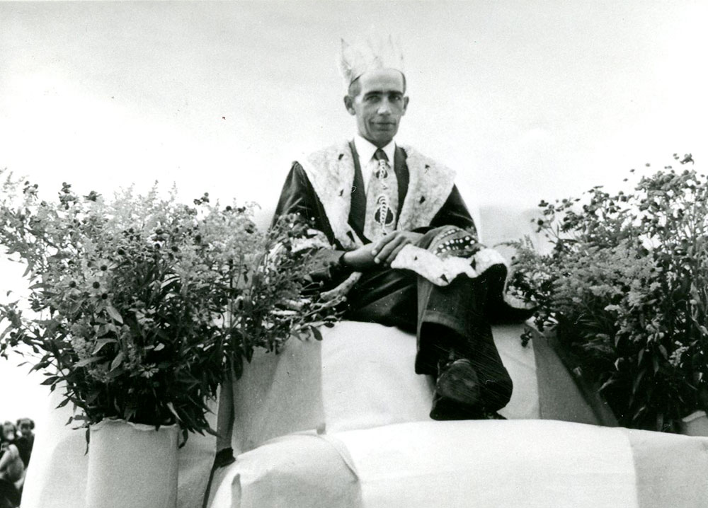 Photo en noir et blanc d'un homme vêtu d'une robe royale et d'une couronne, assis sur un trône et entouré de fleurs. C'est le roi du maïs de 1951.