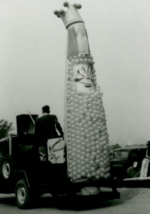 Photo en noir et blanc d’un char de la mascotte du Corn Festival. La mascotte est composée de ballons et porte une couronne.