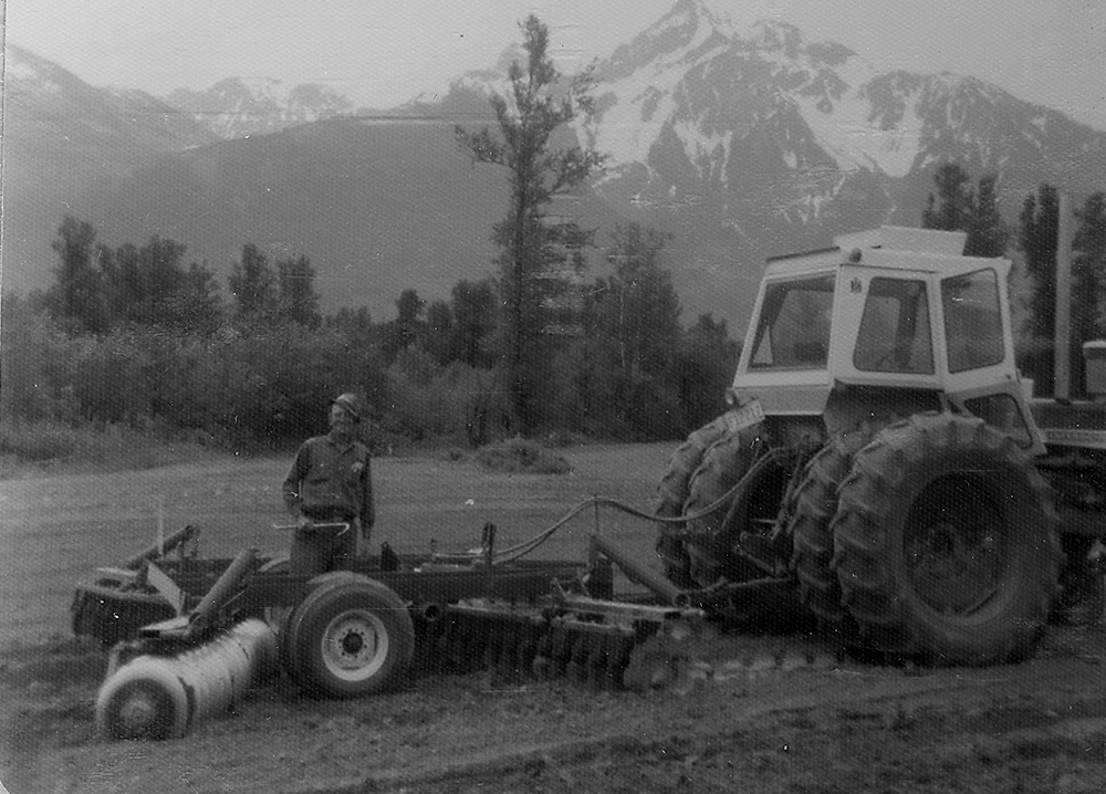 Photo en noir et blanc d'un homme et d'un tracteur tirant une machine à disques dans un champ. Le mont Cheam est en arrière-plan.