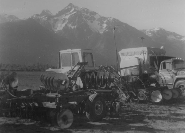 Photo en noir et blanc de deux tracteurs tirant une machine à disques et un camion dans un champ. Le mont Cheam est en arrière-plan.