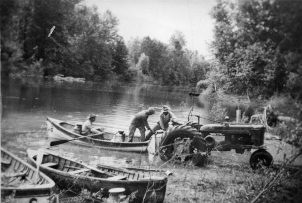 Photo en noir et blanc de trois hommes en train de charger des bidons de lait dans un bateau à rames à partir d'un tracteur.