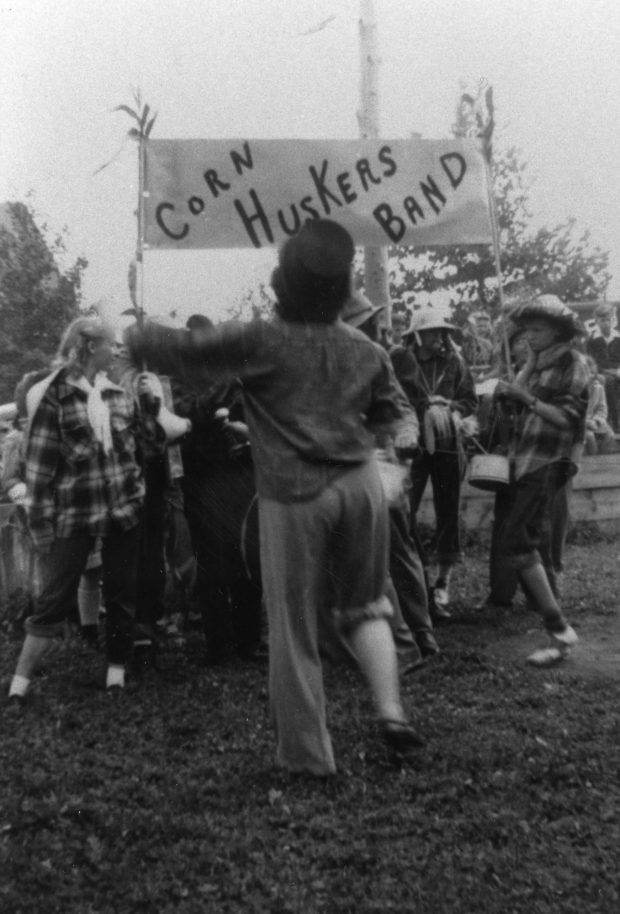 Photo en noir et blanc d’un groupe portant des chemises à carreaux et des chapeaux de paille et portant des instruments de musique. Ils tiennent une pancarte sur laquelle il est écrit « Corn Huskers Band ».