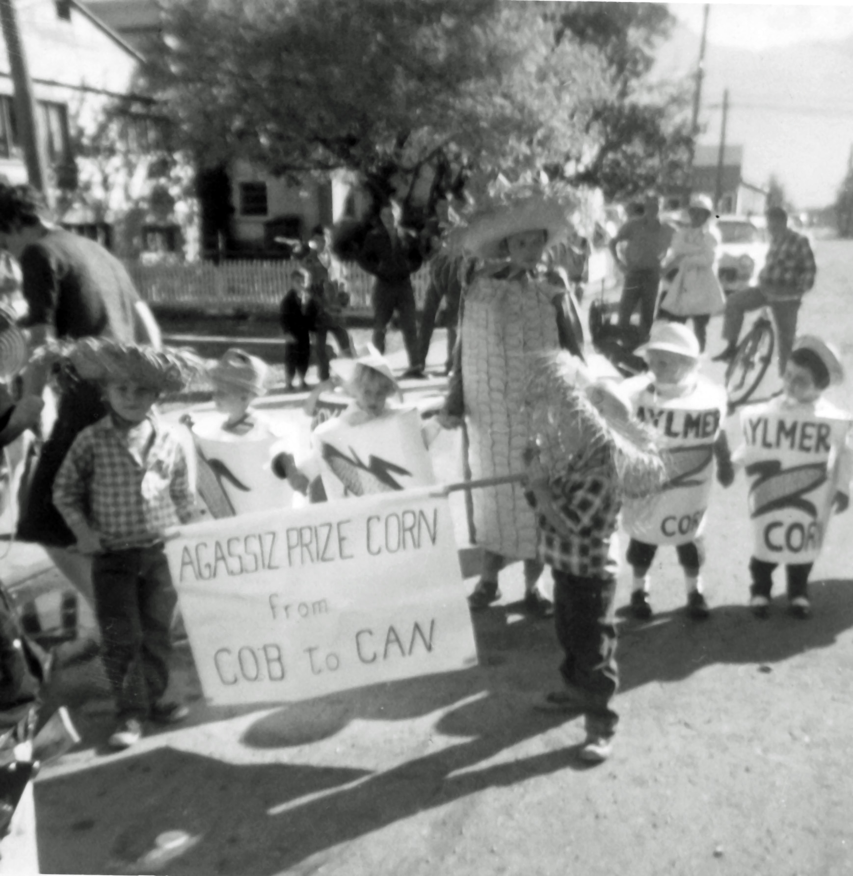Photo en noir et blanc d'enfants dans un défilé, déguisés en épis de maïs et en boîtes de maïs. Deux enfants tiennent une pancarte sur laquelle il est écrit : « Agassiz prize corn from cob to can » (Le maïs primé d'Agassiz, de l'épi à la boîte). 