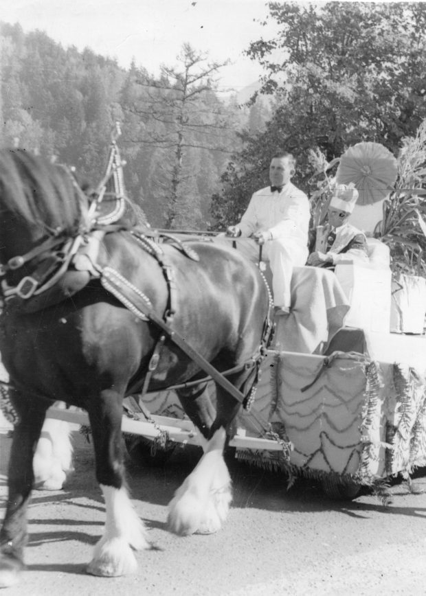 Photo en noir et blanc d'un attelage tirant un char dans un défilé. Le conducteur est vêtu d'un costume blanc et le passager porte une robe royale et une couronne. C’est le roi du maïs de 1951.