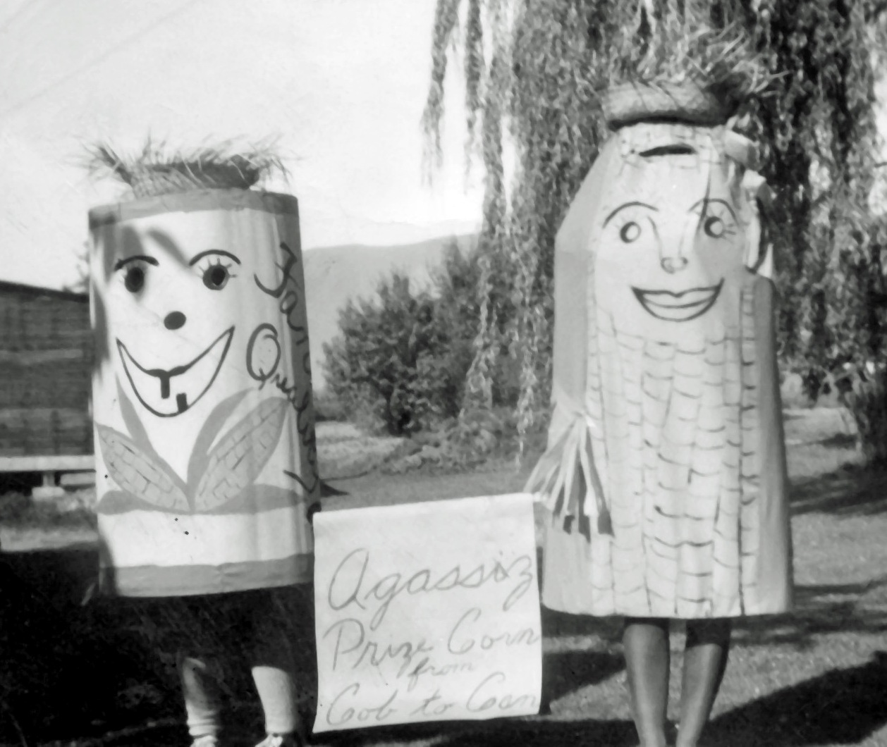 Photo en noir et blanc de deux personnes déguisées en épis et en boîtes de maïs. Ils tiennent une pancarte sur laquelle il est écrit : « Agassiz prize corn from cob to can » (Le maïs primé d'Agassiz, de l'épi à la boîte). 