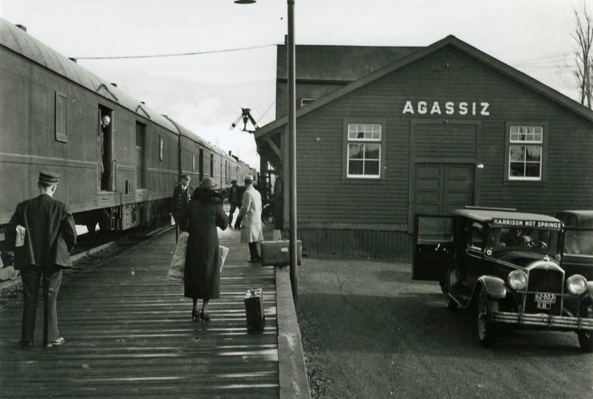 Photo en noir et blanc d'un train à la gare d'Agassiz où des passagers attendent sur un quai. Un taxi de Harrison Hot Springs est au premier plan.