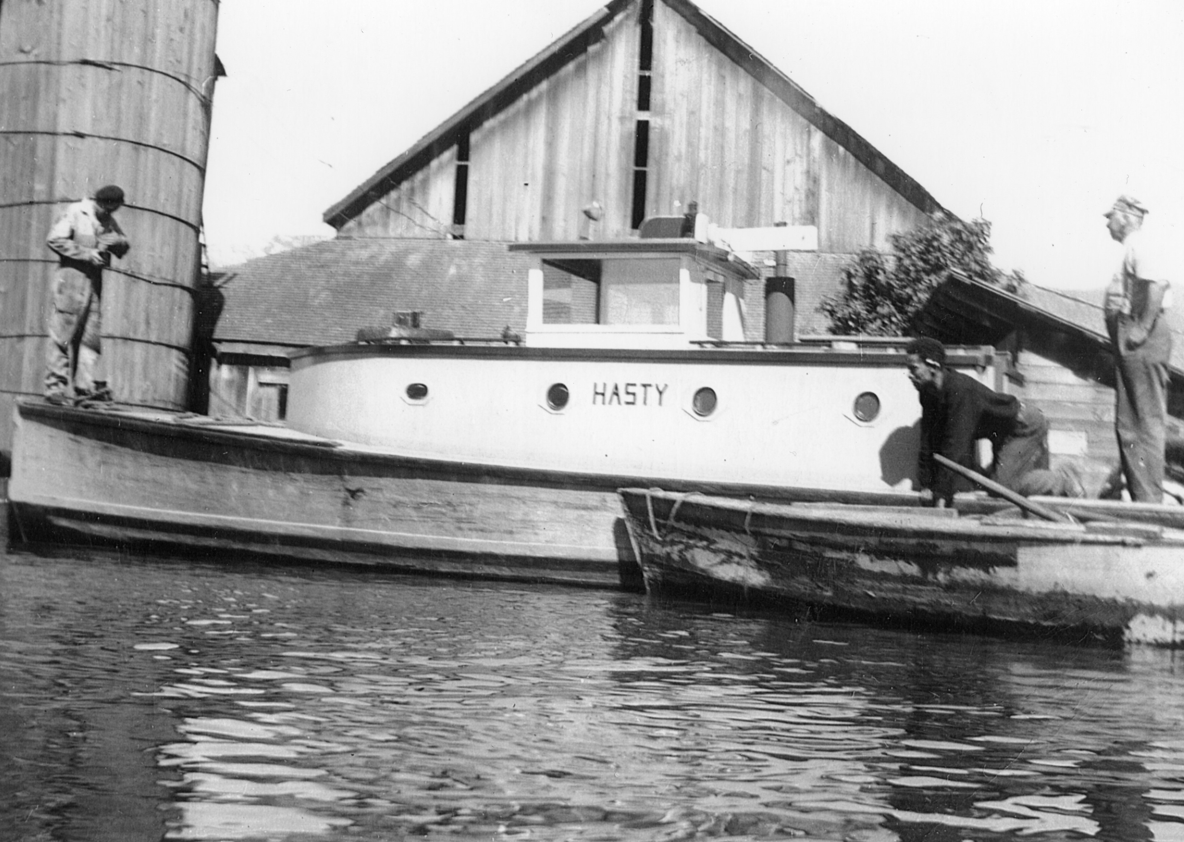 Photo en noir et blanc de trois hommes qui chargent le lait sur le remorqueur « Hasty » devant une grange.