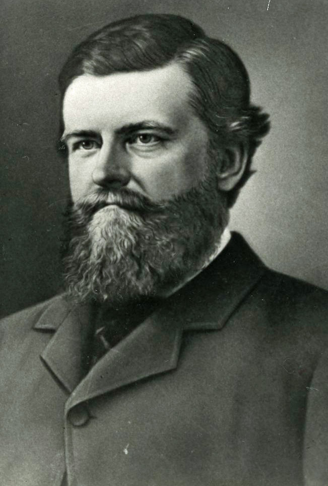 Portrait photographique en noir et blanc d'un homme aux cheveux foncés, portant une barbe et une moustache. Il est vêtu d’un habit avec une cravate.