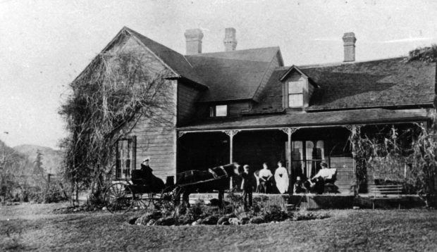 Photo en noir et blanc d’un Homestead. Des femmes sont assises sur la véranda et regardent deux personnes dans une calèche tirée par des chevaux.