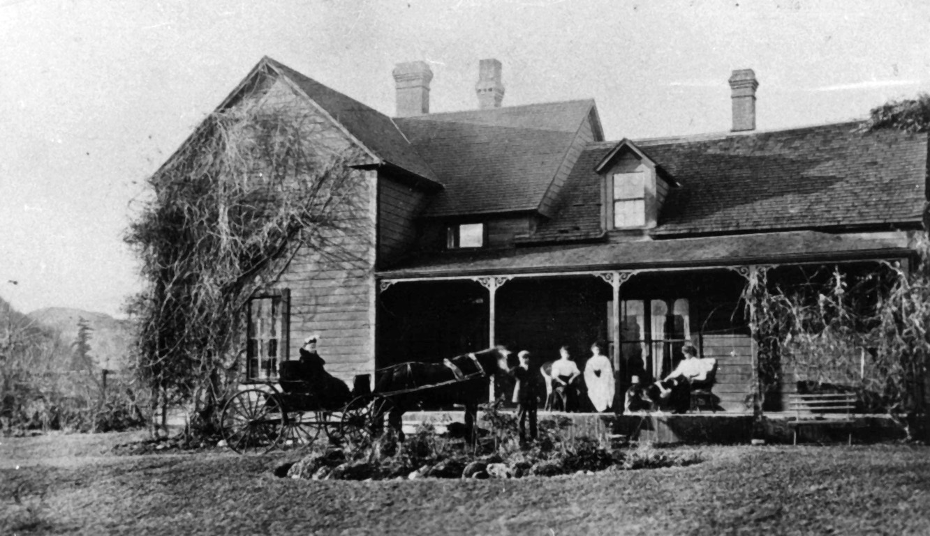 Photo en noir et blanc d’un Homestead. Des femmes sont assises sur la véranda et regardent deux personnes dans une calèche tirée par des chevaux.