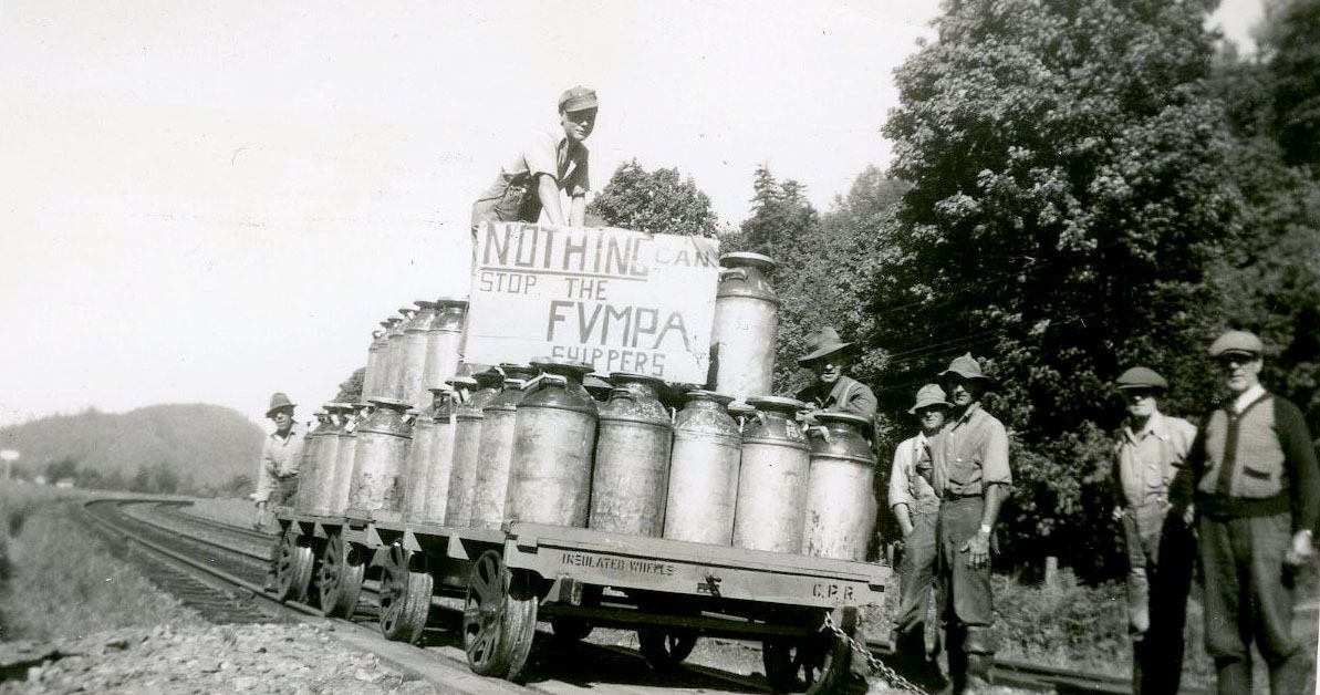 Photo en noir et blanc d'agriculteurs avec un chariot à rails chargé de bidons de lait. Un homme tient un panneau qui dit : « Nothing can stop the FVMPA shippers » (Rien ne peut arrêter les expéditeurs de la FVMPA).