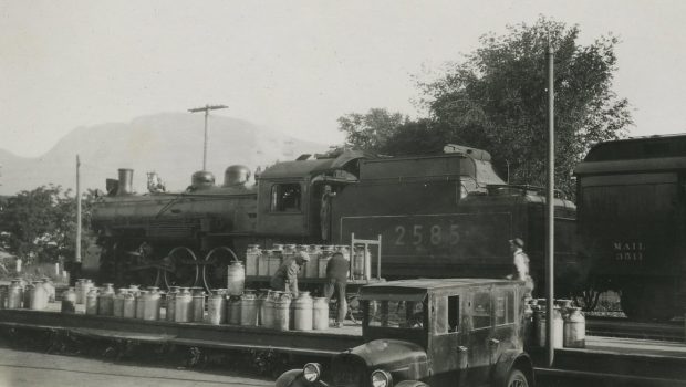 Photo en noir et blanc montrant des hommes chargeant environ 50 grands bidons de lait dans un train. Les bidons sont chargés à partir de la voiture qui se trouve devant.