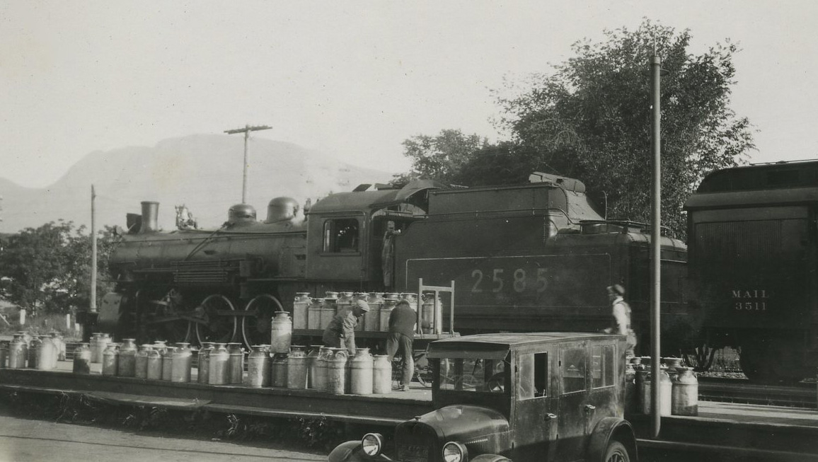 Photo en noir et blanc montrant des hommes chargeant environ 50 grands bidons de lait dans un train. Les bidons sont chargés à partir de la voiture qui se trouve devant.