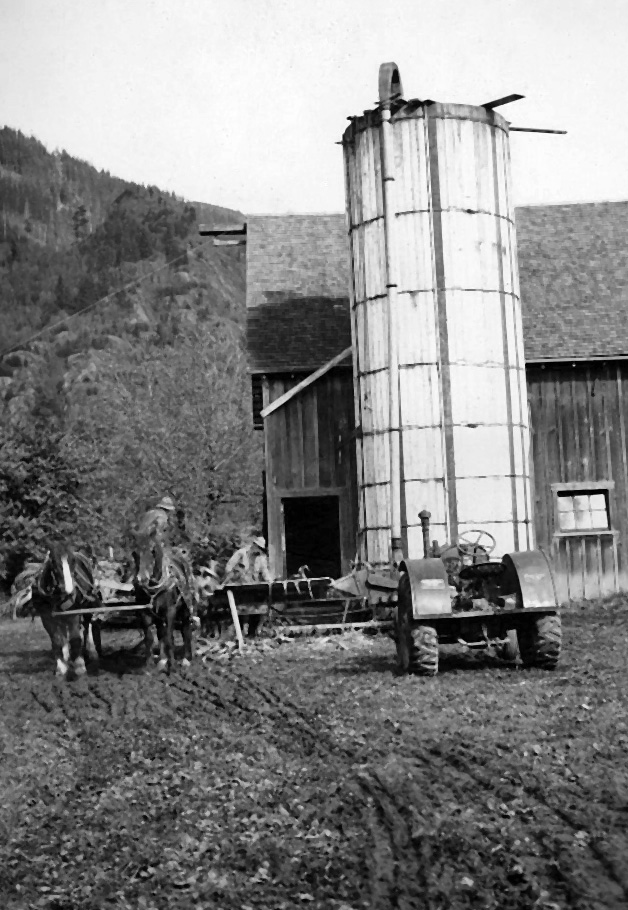 Photo en noir et blanc d'une grange, d'un silo, d'un attelage de chevaux, d'un chariot et d'un tracteur.