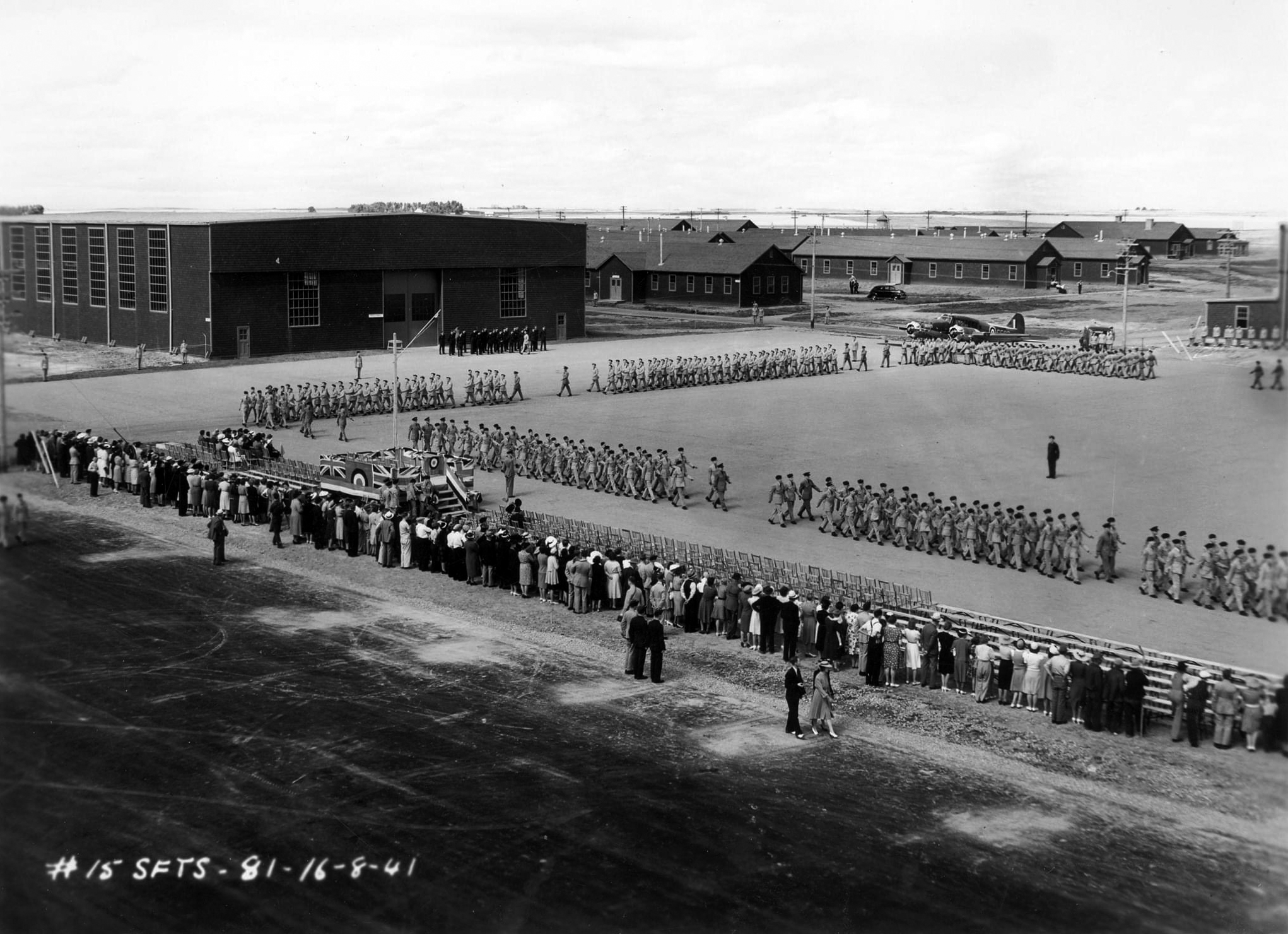 Sept groupes de militaires marchant au pas sur le terrain de parade sous les yeux de la foule