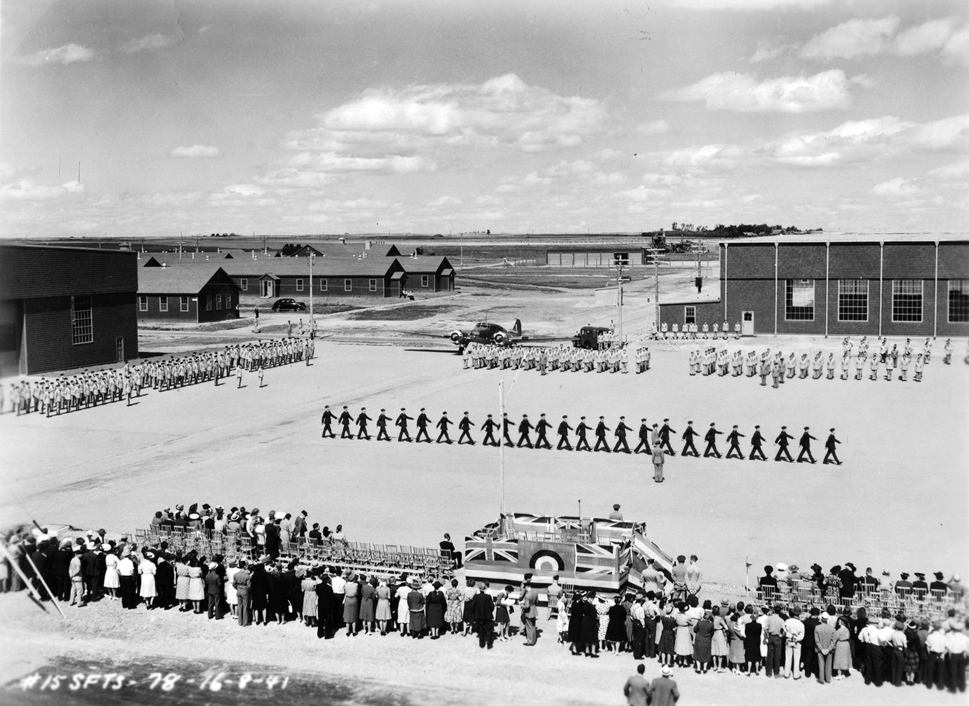 Quatre groupes de militaires marchant au pas sur le terrain de parade sous les yeux de la foule