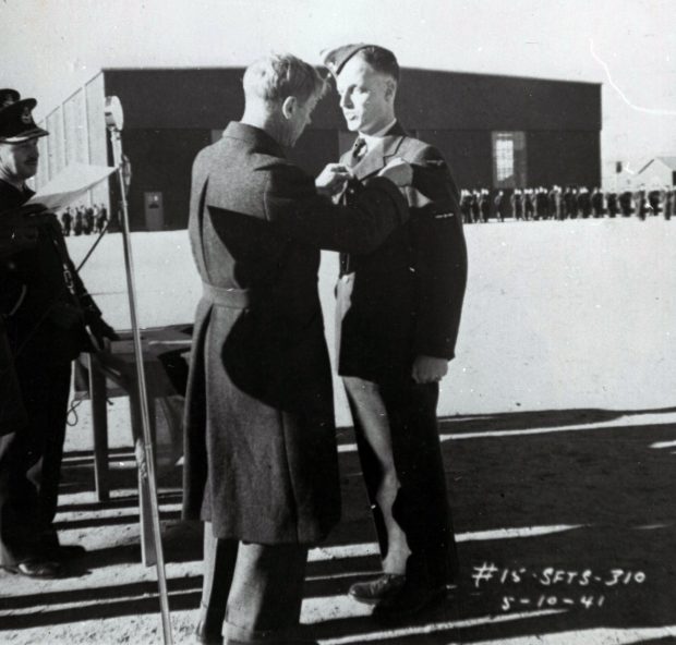 3 hommes, 2 en habit militaire, troupes et hangar en arrière-plan