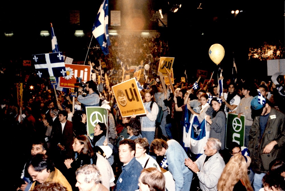 Photographie en couleurs où l’on voit une foule de personnes dans des gradins, portant des drapeaux du Québec et des pancartes.