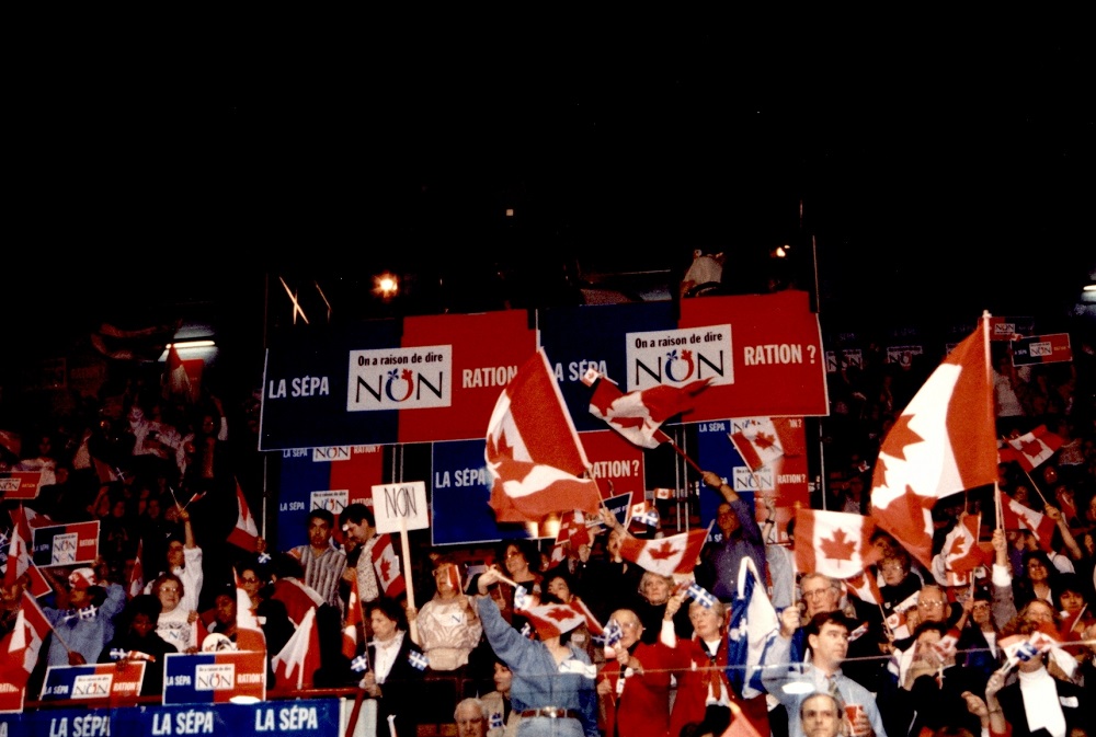 Photographie en couleurs où l'on voit une foule de personnes dans des gradins, portant des drapeaux du Canada et des pancartes.