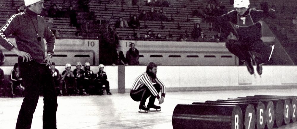 Photographie en noir et blanc où l'on voit trois hommes sur une patinoire. L'un d'eux est en train de sauter par-dessus des barils.