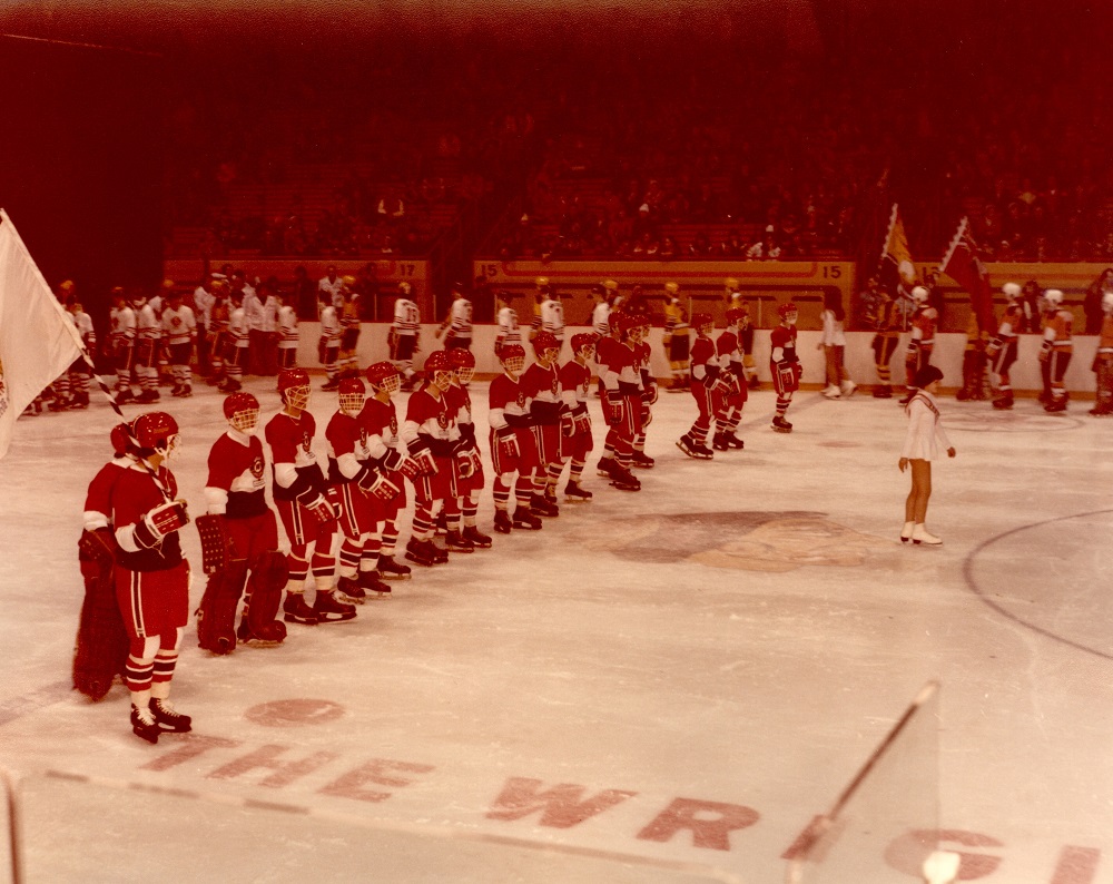 Photographie en couleurs où l’on voit sur une patinoire des dizaines de joueurs de hockey dans leur uniforme. Une jeune fille patine devant les garçons. Des spectateurs se trouvent dans les gradins derrière.