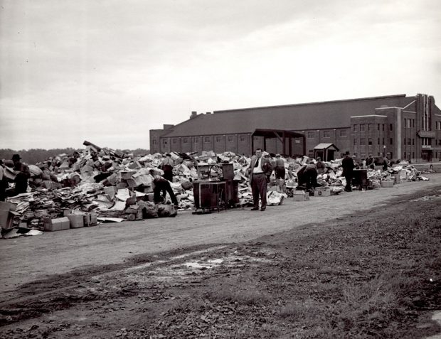 Photographie en noir et blanc montrant un lieu de collecte, situé à côté d’un bâtiment au loin. Une dizaine d’hommes fouillent ce dépôt.