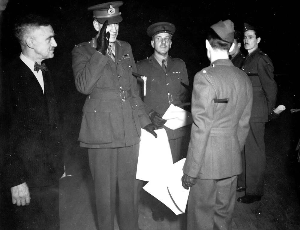 Photographie en noir et blanc où l’on voit quatre militaires en uniforme et un civil. Trois des soldats tiennent à la main des documents, et l’un d'eux effectue un salut militaire.