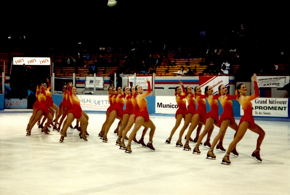Photographie en couleurs où l’on voit 16 femmes en tenue de patinage artistique, en train de faire des mouvements synchronisés sur une patinoire.