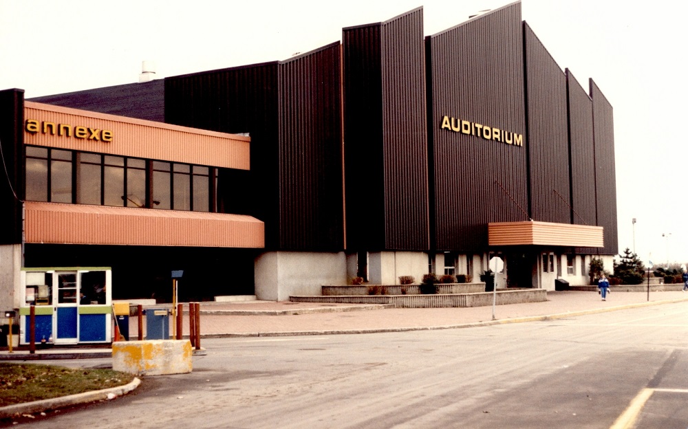 Photographie en couleurs où l’on aperçoit deux bâtiments, l’un avec une devanture en métal noire, et l’autre de couleur orange. Sur la devanture noire est écrit le mot « Auditorium » alors que sur le bâtiment orange est écrit le mot « Annexe ».