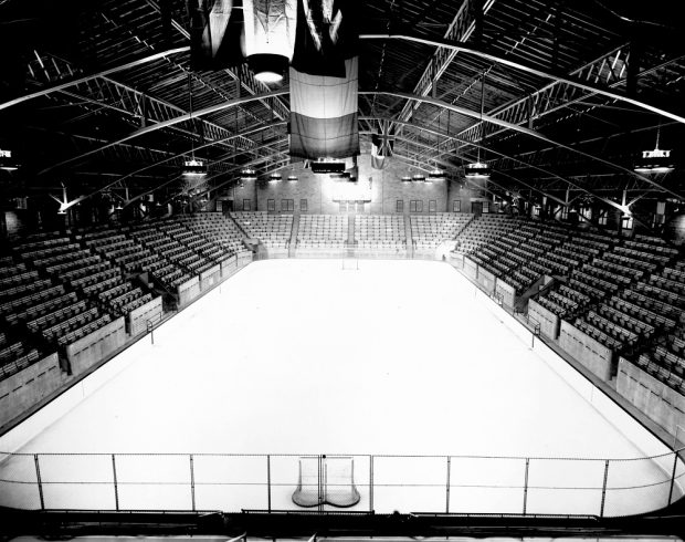 Photographie en noir et blanc de l’intérieur d’un aréna. On y aperçoit une patinoire entourée de gradins vides. Deux filets de hockey se trouvent aux extrémités de la glace et des drapeaux flottent au-dessus de la patinoire.