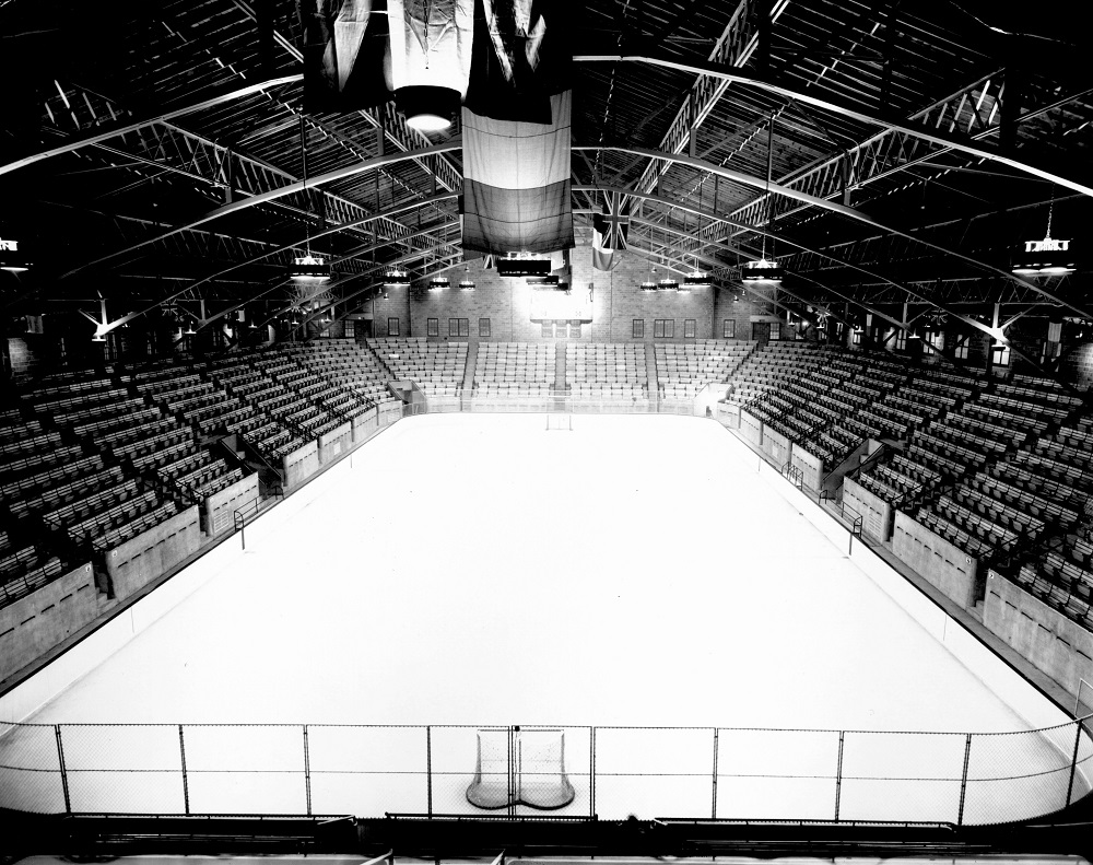 Photographie en noir et blanc de l’intérieur d’un aréna. On y aperçoit une patinoire entourée de gradins vides. Deux filets de hockey se trouvent aux extrémités de la glace et des drapeaux flottent au-dessus de la patinoire.