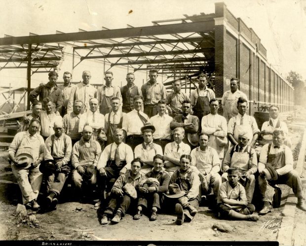 Photographie en noir et blanc montrant un groupe de 33 ouvriers, certains assis, d’autres debout, devant la structure d’acier d’un bâtiment en construction. On ne voit que la structure d’acier et un mur avec un revêtement en briques.