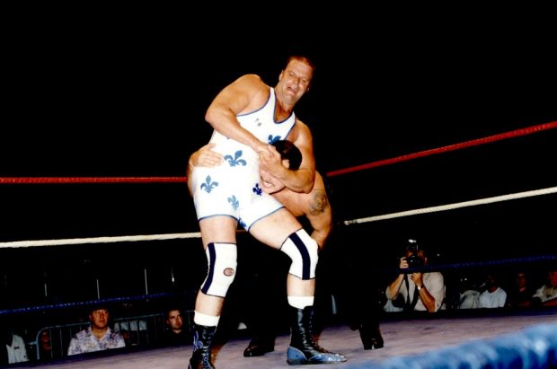 Photographie couleur où l’on voit deux hommes se battre dans une arène. Le lutteur à l’avant-plan porte un costume blanc avec des fleurs de lys bleues et des genouillères.