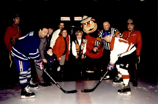 Photographie en couleurs où l’on voit deux joueurs de hockey sur une patinoire en attente d’une mise au jeu. Derrière eux se trouvent cinq personnes sur un tapis : un enfant, un homme en tenue d’arbitre, deux policiers de la Gendarmerie royale du Canada et leur mascotte.