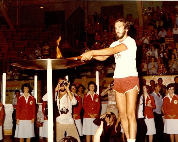 Photographie en couleurs montrant un homme avec des shorts orange et un chandail blanc, qui tient un flambeau allumé au-dessus d’un récipient. Une foule se trouve en arrière-plan.