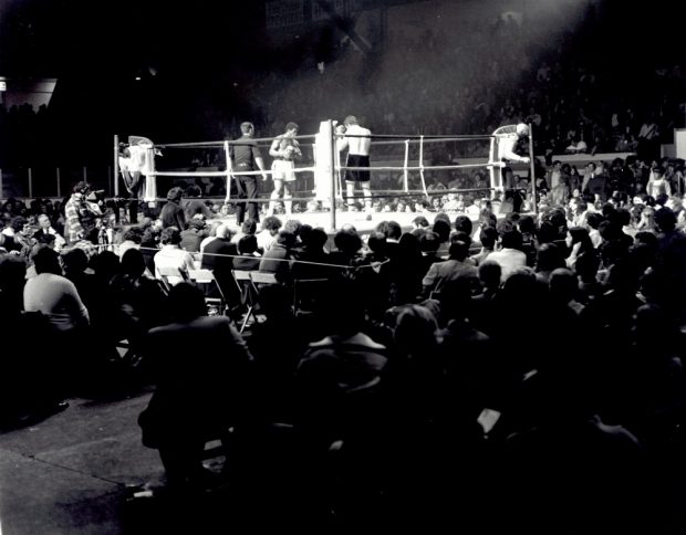 Photographie en noir et blanc où l’on voit deux hommes s’affronter dans une arène de boxe. Une importante foule encourage les deux athlètes.