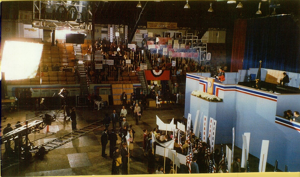 Photographie en couleurs montrant une grande tribune bleue sur laquelle se trouve une personne. Autour de cette tribune se trouvent des personnes tenant des pancartes et des caméras de cinéma.