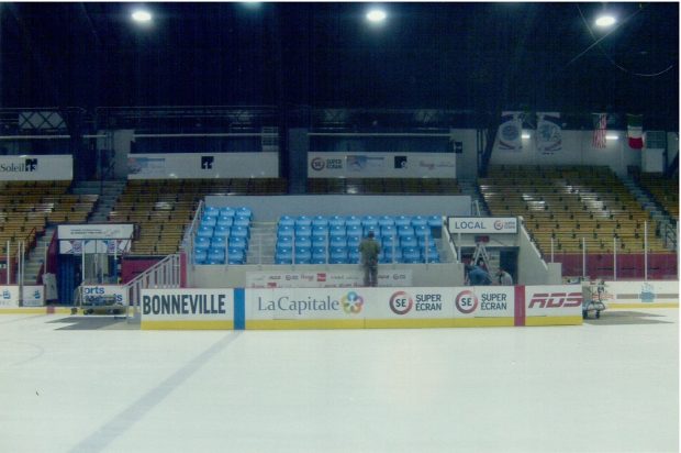 Photographie en couleurs où l’on voit à l’avant une patinoire et sa bande protectrice, sur laquelle se trouvent plusieurs noms et logos de compagnies. Au centre se trouvent plusieurs rangées de bancs de couleur bleue. Des gradins bruns et jaunes les entourent.